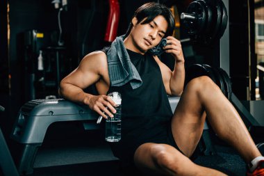 Spor kıyafetleriyle eğitilmiş Asyalı bir gencin portresi spor salonunda antrenman, ağırlık eğitimi, sağlıklı yaşam tarzı konsepti sonrasında elinde su ya da proteinle bankta dinleniyor.