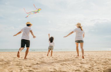 Asyalı bir aile sahilde koşarken gökyüzünde uçurtma uçuruyor. Mutlu ebeveynler, anne ve babalar ve çocukları uçurtmayla oynuyorlar, aileler birlikte vakit geçiriyorlar, yaz günü.
