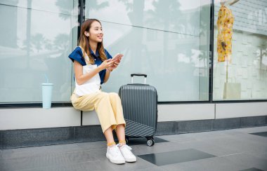 Elinde bavuluyla oturan şık bir kadın, tren istasyonunda ulaşımı beklerken cep telefonunu bağlı kalmak ve birden fazla iş yapmak için kullanıyor. O verimli ve modern..