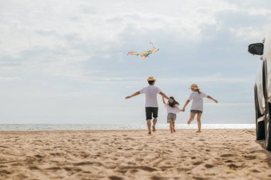 Asyalı bir aile sahilde koşarken uçurtma uçuruyor. Mutlu anne ve baba ve çocukları uçurtmayla oynarken araba yakınlarda park edilmiş ve aile birlikte vakit geçiriyorlar.