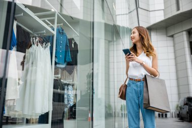 Alışveriş çantalı güzel bir kadın, cep telefonunu karıştırıyor, bir moda mağazasının önünde duruyor. Modern teknolojinin rahatlığı ve tüketim zevkinin tadını çıkarıyor..