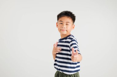 Neşeli Asyalı genç çocuk kameraya iki silah parmağı hareketi yapıyor. Dost canlısı ve küstah bir ifadeyle. Beyaz arka planda, birincil çocuk pozitif ve neşe yayar.