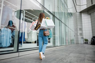 Elinde alışveriş torbaları olan şık bir kadın yaz sezonunda giyim mağazasının önünde yürüyor. O bir moda tutkunu, her zaman en son moda kıyafetleri ve aksesuarları arar.