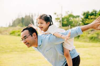 Baharda çimenlerde oynayan Asyalı bir aile mutlu anne-baba, baba-kız babaya biniyor açık havada eğleniyor, baba kızını taşıyor, aile ferahlıyor.