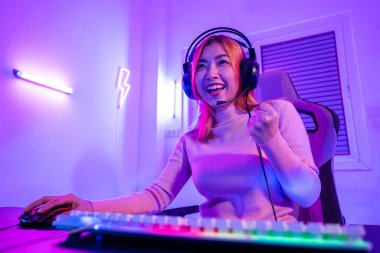 Zafer kazanmak. Asyalı oyuncu online video oyunu oynuyor. Masaüstü bilgisayarında geceleri mor ışık yanıyor. Oyun kulaklıklı mutlu kadın başarılı olduğu bilgisayarı kullanıyor, E-Sport kavramı.