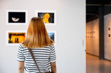 Asyalı kadın renkli çerçeveli resimlerin önünde dikilip beyaz duvara bakıyor, insanlar fotoğraf çerçevesinde sergi sanat galerisine yaslanıyor,