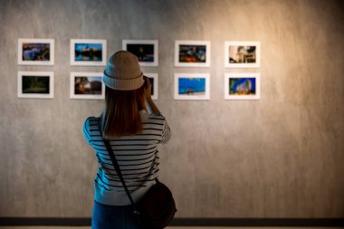 Asyalı kadın, çerçeveli resimlerin önünde resim galerisi koleksiyonu çekiyor beyaz duvarda kameralı resimler, fotoğraf çerçevesinde fotoğrafçı saati sergi sanat galerisine yaslanıyor.