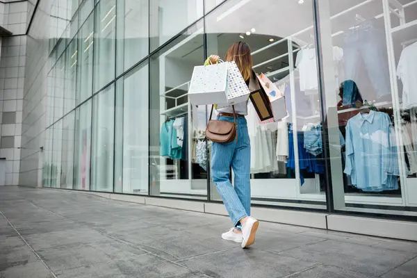Kalabalık bir kalabalık ve hareketli şehir mimarisi tarafından çevrili alışveriş torbalarıyla mağazanın önünde yürüyen bir kadın. Şık alışverişçi, her zaman en son trendleri arıyor.