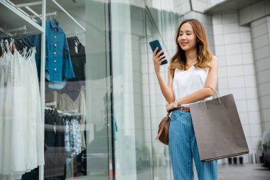 Çantalı ve akıllı telefonlu mutlu bir kadın güneşli bir günde birden fazla iş yapıyor. Manikinlerle bir moda mağazasının önünde dikilirken internette geziniyor..