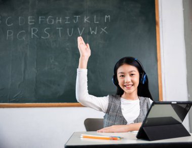 Genç bir kız sınıftaki bir masada oturuyor, elini kaldırıyor ve onu havaya kaldırıyor. Kulaklık takıyor ve tablet ya da laptop kullanıyor.