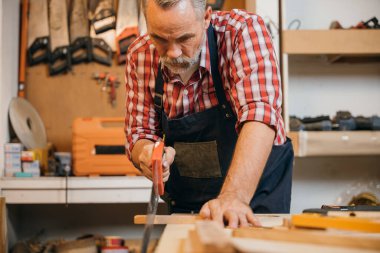 Kırmızı ekose gömlekli ve mavi tulumlu bir marangoz tezgahın üzerinde el testeresiyle bir tahta parçasını testereyle kesiyor, marangozluk projesinde kesin kesikler üzerine yoğunlaşıyor, Ulusal Marangozlar Günü.