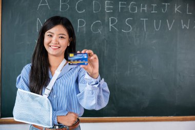 Üzerinde alfabe olan karatahtanın önünde kredi kartı tutan kadın. O gülümsüyor. Kolum kırıldı. Öğretmenin kendine güveni kazadan sonra kırılmış ve tedavi için kol bandı takıyor, destek kolu takıyor.