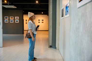 Asyalı bir kadın sanat galerisinde tablet tutuyor. Çerçeveli resimlerin önünde beyaz duvarda, fotoğraf çerçevesinde genç bir kadın sergi sanat galerisinde dijital kitap tutuyor.