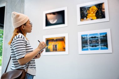 Asyalı kadın sanat galerisindeki tablete bilgi not alıyor. Çerçeveli resimlerin önünde duvarlara bakıyor. Fotoğraf çerçevesindeki genç kişi sergi sanat galerisinde dijital kitap yazıyor.