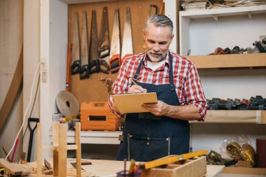 Ekose gömlekli ve tulumlu bir adam atölyesinde ahşap bir proje üzerinde çalışıyor. Kalemle panoya resim çiziyor. Aletler ve ahşap ile çevrili, marangozluk ve marangozluk, Ulusal Marangozlar Günü