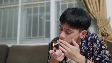 Asyalı genç adam kanepede oturup sigara içerken evde dinleniyor. Erkekler kanepede sigara içerken, arka planda bir gitar var.