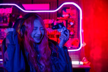 Kazanmak. Happy Gamer sanal turnuva için oyun kumandası kullanıyor bilgisayarla online video oyunu oynuyor kutlama yapmak için elini kaldırıyor oyun kulaklığı takan Asyalı kadın heyecanlanıyor