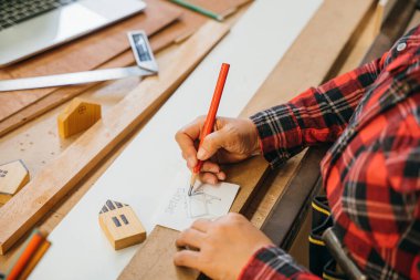 Kırmızı ekose gömlekli biri kırmızı kalemle çizim masası planları yapıyor. Tahta tahtalar ve minyatür evlerle çevrili. Mimarlık, inşaat ve DIY proje kavramları için mükemmel