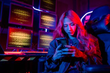 Bilgisayar ekranının önündeki Asyalı kadın cep telefonu, neon ışık, Hacker sistem kontrol odasında siyah ceket giyiyor güvenlik görevlisi birden fazla monitörde birlikte çalışıyor akıllı telefon.