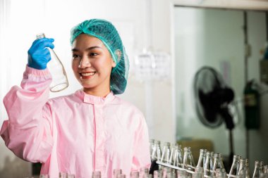 Kimya bilimi işçisi üniformalı bir QC kadın titizlikle cam şişeleri inceliyor meyve suyu kalitesinde içki fabrikası endüstrisinde yüksek standartları güvence altına alan.