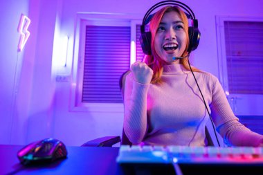 Zafer kazanmak. Asyalı oyuncu online video oyunu oynuyor. Masaüstü bilgisayarında geceleri mor ışık yanıyor. Oyun kulaklıklı mutlu kadın başarılı olduğu bilgisayarı kullanıyor, E-Sport kavramı.