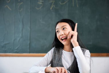 Sınıfta bir kız oturuyor ve parmağını işaret ediyor. Gülümsüyor ve tahtaya bakıyor.
