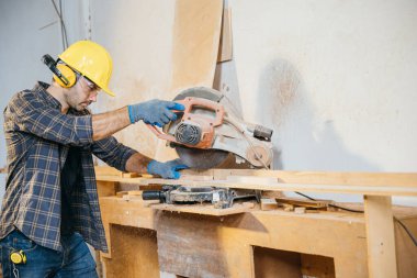 Odun kesmek için testere kullanan sarı şapkalı bir adam. İnşaat, ev tadilatı ve ahşap işçiliği için ideal elektrikli aletlerle kusursuz kesim teknikleri, Ulusal Marangozlar Günü