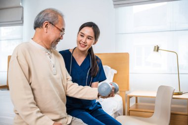 Genç hemşire fizyoterapisti, yaşlı bir adama evde halter kaldırmada yardım ediyor. Asyalı hemşire, yaşlı emekliliğe destek oluyor. Koltukta otururken halterli egzersiz, fiziksel tedavi, sağlık konsepti kullanıyor.