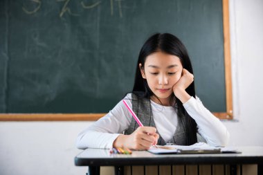 Bir kız masada oturuyor ve pembe bir kalemle yazıyor. Beyaz bir gömlek ve gri bir yelek giyiyor. Oda bir karatahta ve karatahta ile süslenmiş.