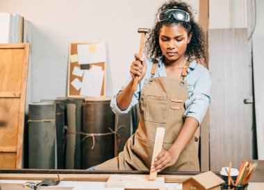 Afro-Amerikalı marangoz kadın bir marangoz dükkanında bir sandalye için bir tahta parçasını çekiçliyor. Ahşap mobilya ustalığı, atölyede zanaat, DIY, Ulusal Marangozlar Günü