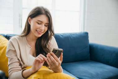 Kanepede oturan gülümseyen bir kadın telefonunu tutar ve mesaj yazar. Evdeki rahatlık ortamında, iletişim ve teknoloji sayesinde rahatlıyor. Kadın dinleniyor..