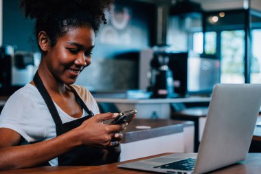 Kafe sahibinin portresi. Önlüklü, akıllı telefon kullanan siyahi bir kadın. Çok görevli barista iletişimi yönetir. Kafesi mükemmel hizmet verir..