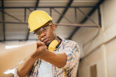 Afro-Amerikan marangoz atölyede tahta taşırken bir tahta parçasını teftiş ediyor. İnşaat işleri, ahşap kalite teftişi ve marangozluk, Ulusal Marangozlar Günü.