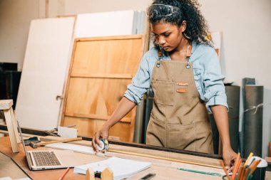 Afro-Amerikalı marangoz kadın, mobilya odununu mezurayla ölçüyor. Bir atölyede ahşap bir kalas üzerinde çalışmak, işçilik ve DIY projeleri ile çalışma tezgahında aletler, Ulusal Marangozlar Günü