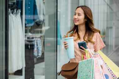 E-ticaret ve tüketim kavramı: akıllı telefonunu kullanan ve bir mağazanın vitrininde çanta tutan genç bir kadın..