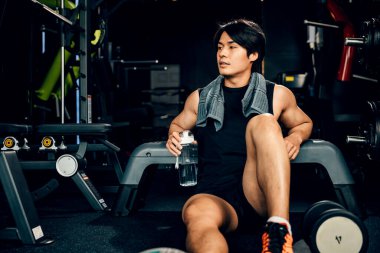 Spor kıyafetleriyle eğitilmiş Asyalı bir gencin portresi spor salonunda antrenman, ağırlık eğitimi, sağlıklı yaşam tarzı konsepti sonrasında elinde su ya da proteinle bankta dinleniyor.