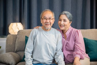 Mutlu Asyalı yaşlı çiftin portresi rahat bir koltukta otururken birini selamlıyor, yaşlı adam ve kadın evdeki bir kanepede yan yana oturuyor.