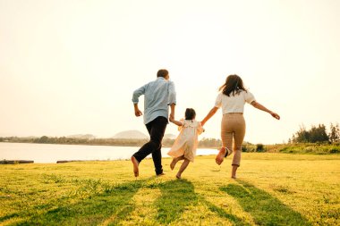 Asyalı bir aile eğleniyor ve doğanın tadını çıkarıyor. Güneşli bir günde yeşil bir bahçede el ele koşup oynuyorlar. Mutlu bir aile günü.