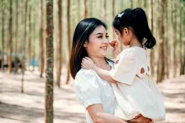 Asyalı bir anne, huzurlu bir orman ortamında kızını kucağında taşıyor, mutlu bir gülümseme anne ve küçük çocuk, dışarıda birlikte oynuyorlar, tatilde aile günü anıları.