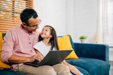 Gülümseyen Asyalı bir baba, modern oturma odalarında e-öğrenim için kızıyla birlikte evde çalışıyor. Bağlayıcı mutlulukları ve birliktelikleri odak noktasında..