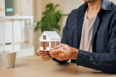 Emlakçının elinde bir minyatür ev modeli var. Mülk, finans, ev kredisi ve konut kavramlarını temsil ediyor. Bir ajanın rüya ev ve kredi çözümlerini güvence altına almasındaki rolünü yansıtır..