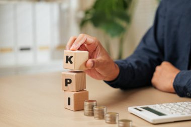 KPI 'yi madeni paraların ve hesap makinesinin yanında yazan tahta küp blokları tutan bir iş adamının yakın çekimi. Anahtar Performans İndeksi planlama, çalışan performans takibi ve iş mali analizini temsil eder.