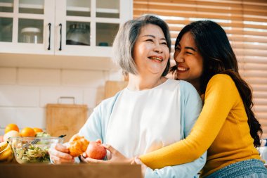 Önlük giymiş genç bir Asyalı kadın ve yaşlı annesi mutfakta yiyecek ve içeceklerle kaynaşıyorlar. Neşeli ifadeleri aile birlikteliğinin ve eğlencenin neşesini gözler önüne seriyor..