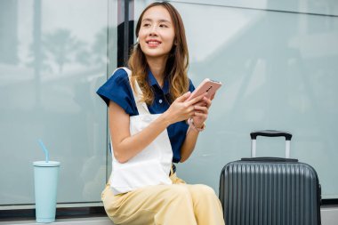 Genç bir kadın bavuluyla oturup rahatlıyor, cep telefonunu sosyal medyada gezinmek ve havaalanında uçağını beklerken bağlı kalmak için kullanıyor. O modaya uygun ve modern..