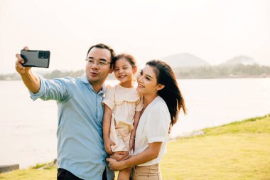 Asyalı bir aile akıllı telefondan doğa parkında fotoğraf çekiyor, mutlu bir gülümseme baba ve kız çocuğu cep telefonuyla fotoğraf çekiyor, turist hayatı tarzı aktiviteler yapıyor.