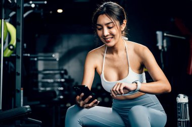 Asyalı bir kadın spor salonunda egzersiz yaptıktan sonra, spor salonunda dinleniyor ve tüm güncellemeleri kontrol ediyor. Kadın cep telefonu ve spor salonunda spor yaparken uygulanan akıllı saat uygulaması, sağlıklı yaşam tarzı.