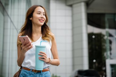 Beyaz tişörtlü şık bir kadın akıllı telefonunu kullanarak şehir sokaklarında bir bardaktan kahve yudumluyor..