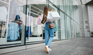 Kalabalık bir caddede alışveriş çantalarıyla gezen genç bir kadın, sevimli bir vitrinle bir giyim mağazasının önünde. Alışverişkoliktir, her zaman en iyi moda anlaşmalarını arar..