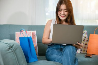 Mutlu kadın müşteri web sitesinden satın almaktan memnun, Asyalı genç bayan alışveriş torbalarıyla kanepede oturuyor bilgisayarla evdeki oturma odasında online alışveriş yapıyor.