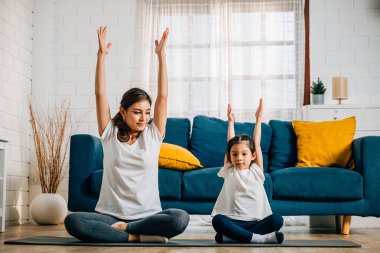 Bir anne ve kızı, esneme ve denge üzerine odaklanmış iç açıcı bir aile yogası seansını paylaşıyorlar. Gülümsemeleri, evde beraberlik ve mutluluk göstergesidir..